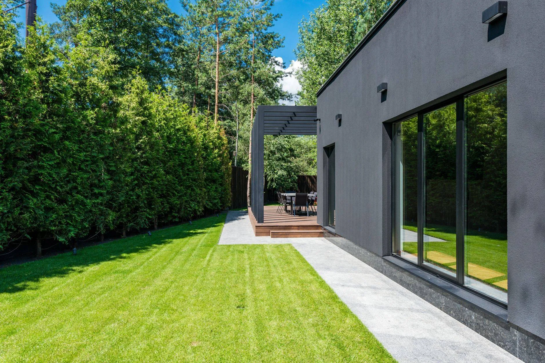 A striped lawn beside a grey contemporary house, a tall hedge along the boundary