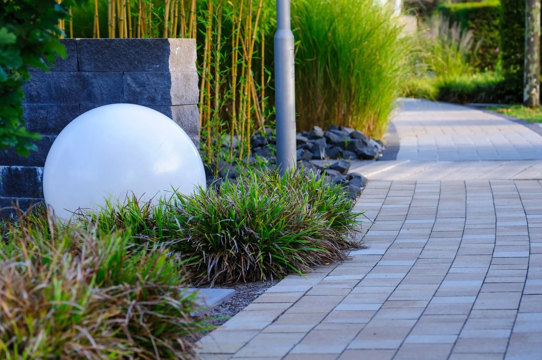 A curved block-paved path with a globe light beside grasses and bamboo